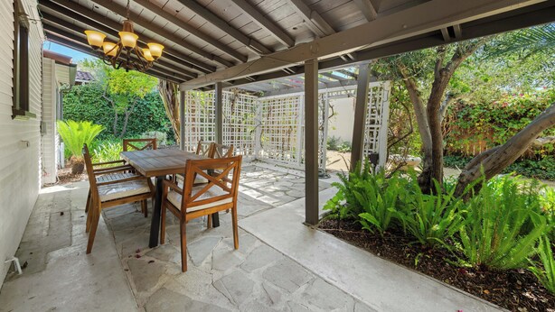 Un patio cubierto con mesa y sillas de madera, rodeado de follaje y una valla blanca.