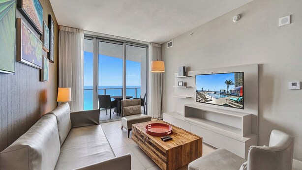 A modern living room with a sofa, coffee table, and TV. There is a balcony with a view of the sea. - Best View in Lyfe Hollywood Beach (Hollywood)