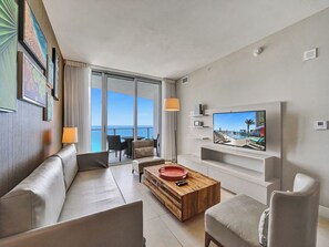 Ein modernes Wohnzimmer mit Sofa, Couchtisch und Fernseher. Ein Balkon bietet Blick aufs Meer.