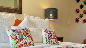 A bed with white linens and colorful embroidered pillows, a black lamp, and heart-shaped wall decorations.
