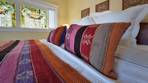 Una cama con almohadillas de colores a rayas y una colcha blanca, una ventana con vitrales y una pared con obras de arte enmarcadas.