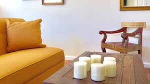 Una sala de estar con un sofá color mostaza, una mesa de centro de madera con velas, una silla de madera y dos cuadros en la pared.