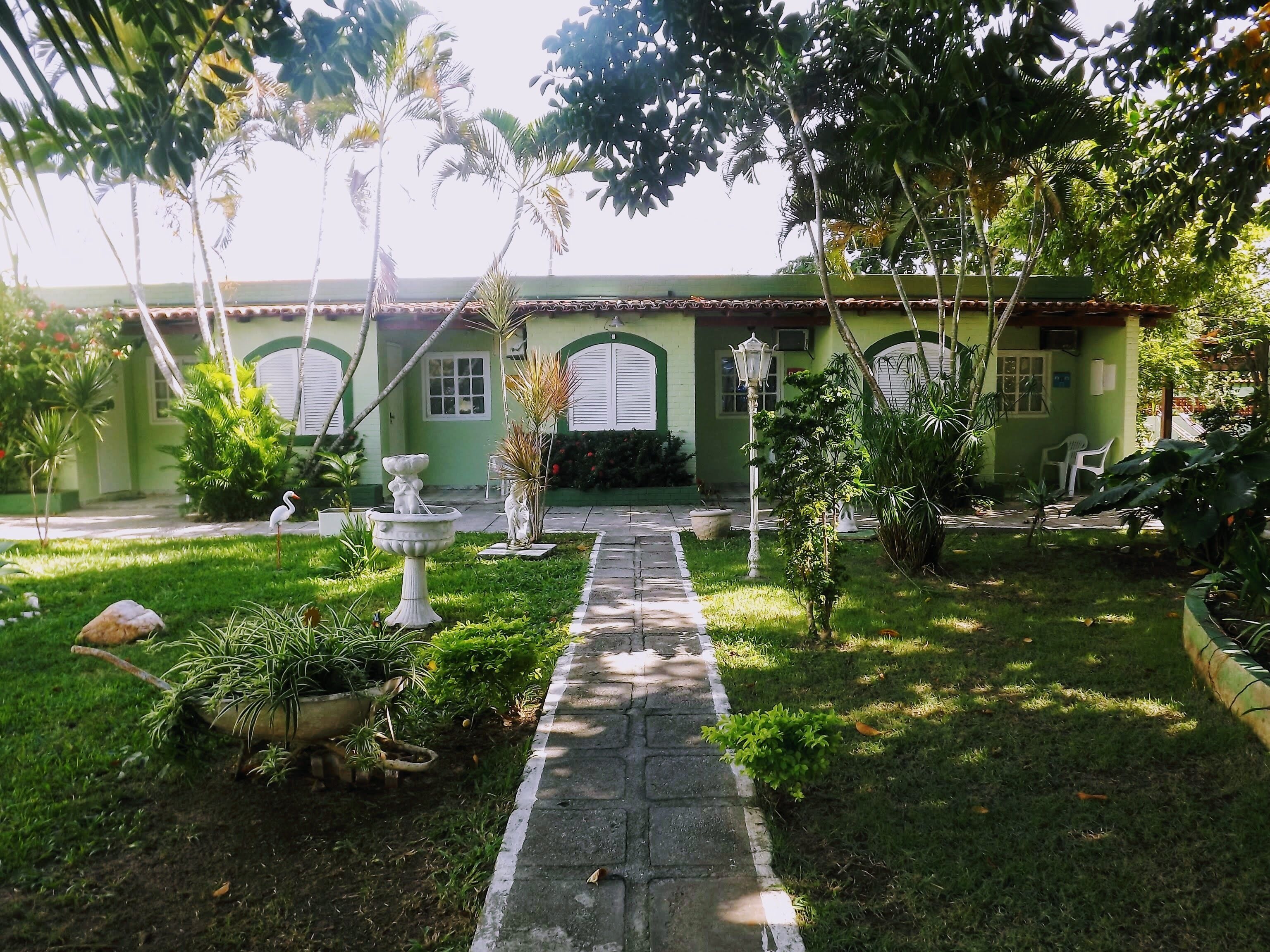 A courtyard with a pathway leading to a building with green walls and arched windows. There are potted plants, a birdbath, and a white lamp post.