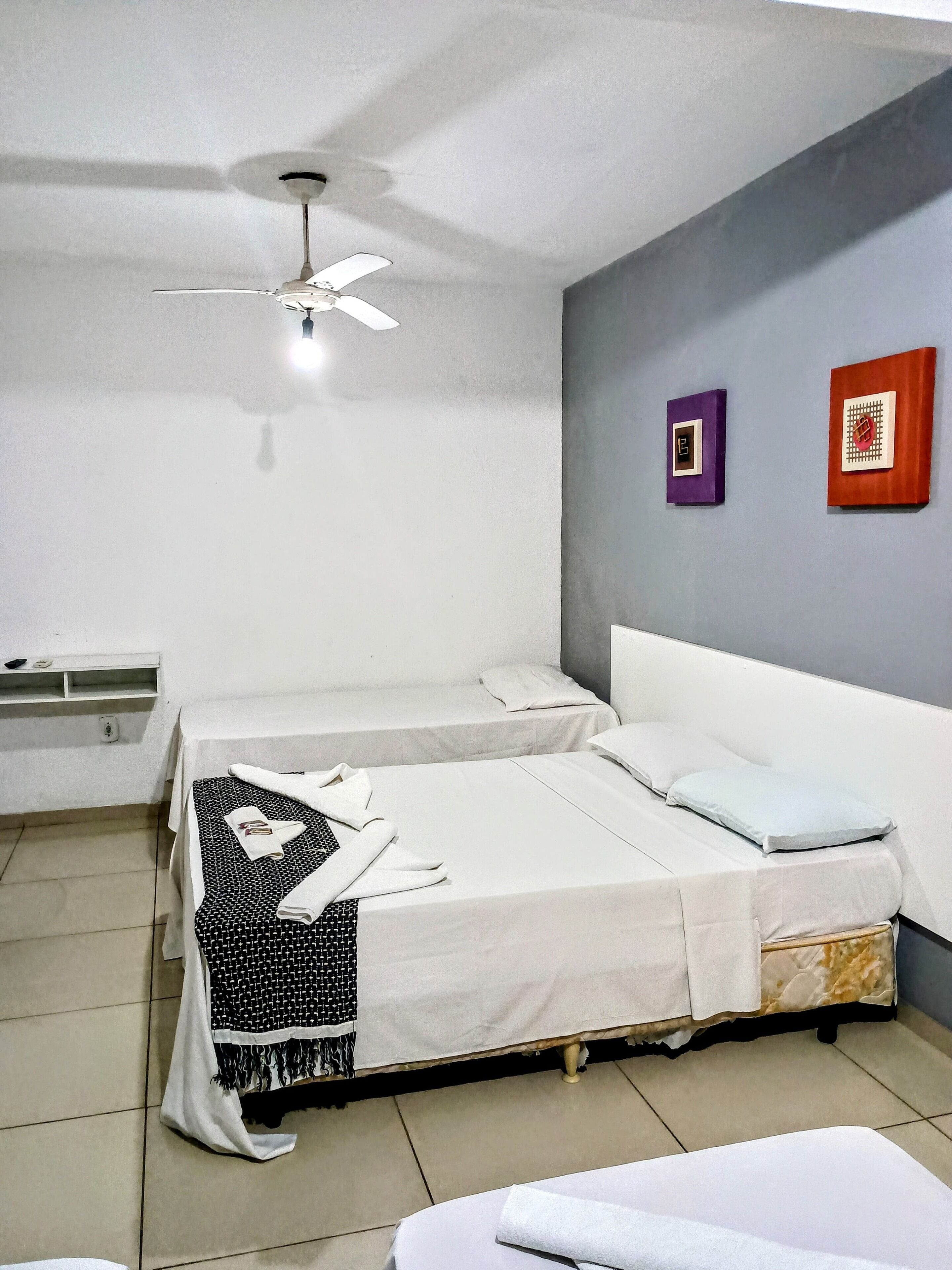 A hotel room with two beds, a ceiling fan, a small shelf, and two wall-mounted clocks.