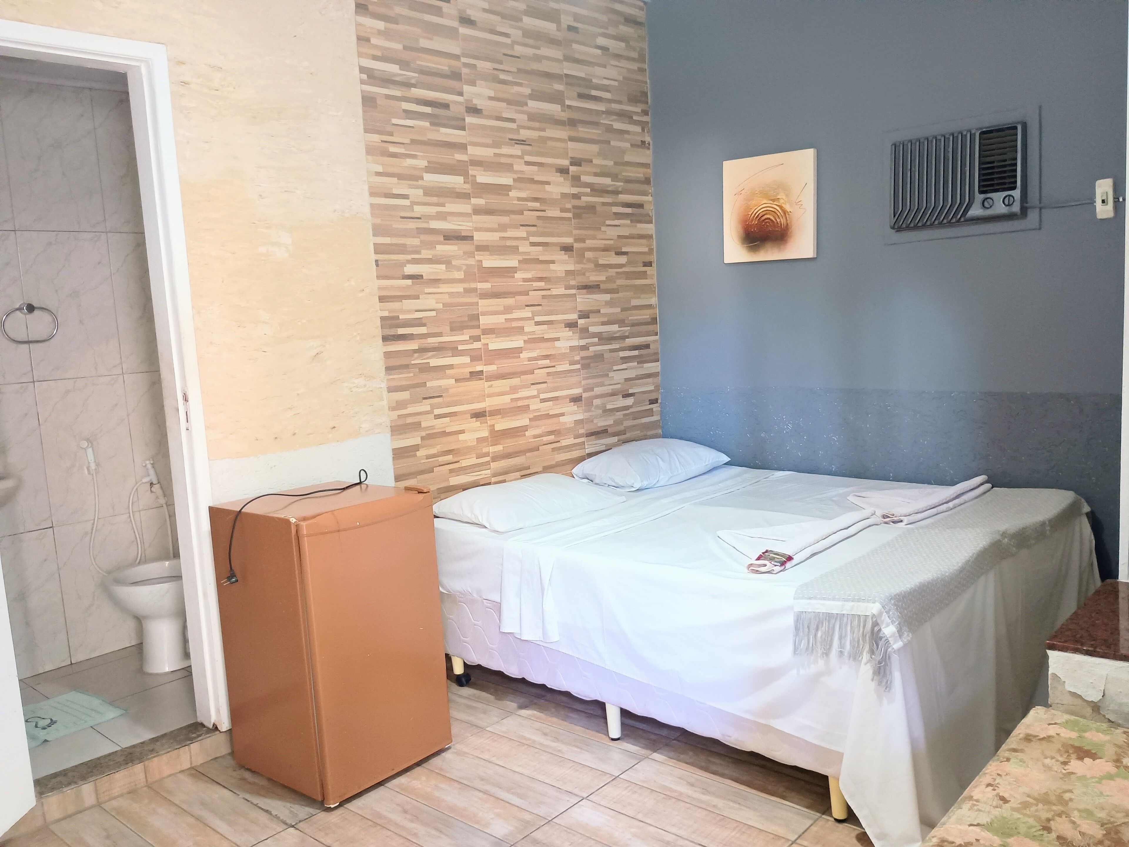 Een slaapkamer met een bed, een houten hoofdbord, een luchtventilator aan de muur en een klein bruin kastje.