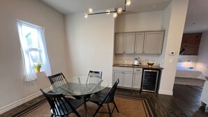 A dining area with a glass table and black chairs, a kitchen with white cabinets, and a wooden floor.