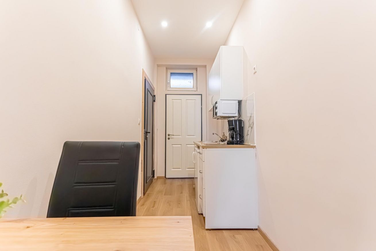 A narrow hallway with a dining table and chairs, a small kitchenette with a refrigerator, and a door leading to another room.