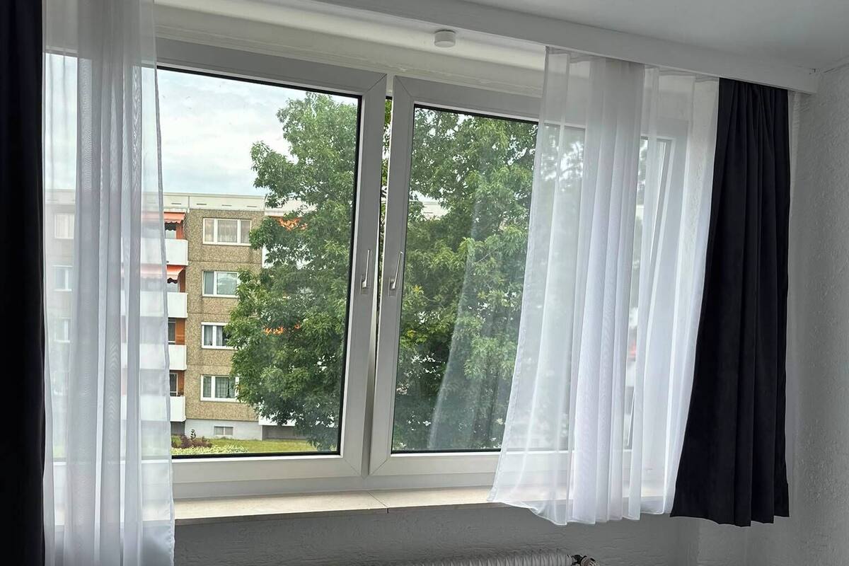 A hotel room with a bed, a chair, a radiator, and a window with curtains.