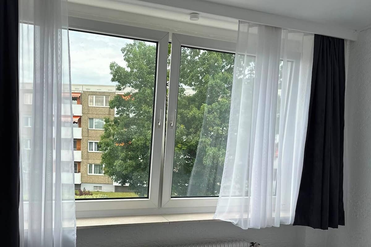 A hotel room with a bed, a chair, a radiator, and a window with curtains.
