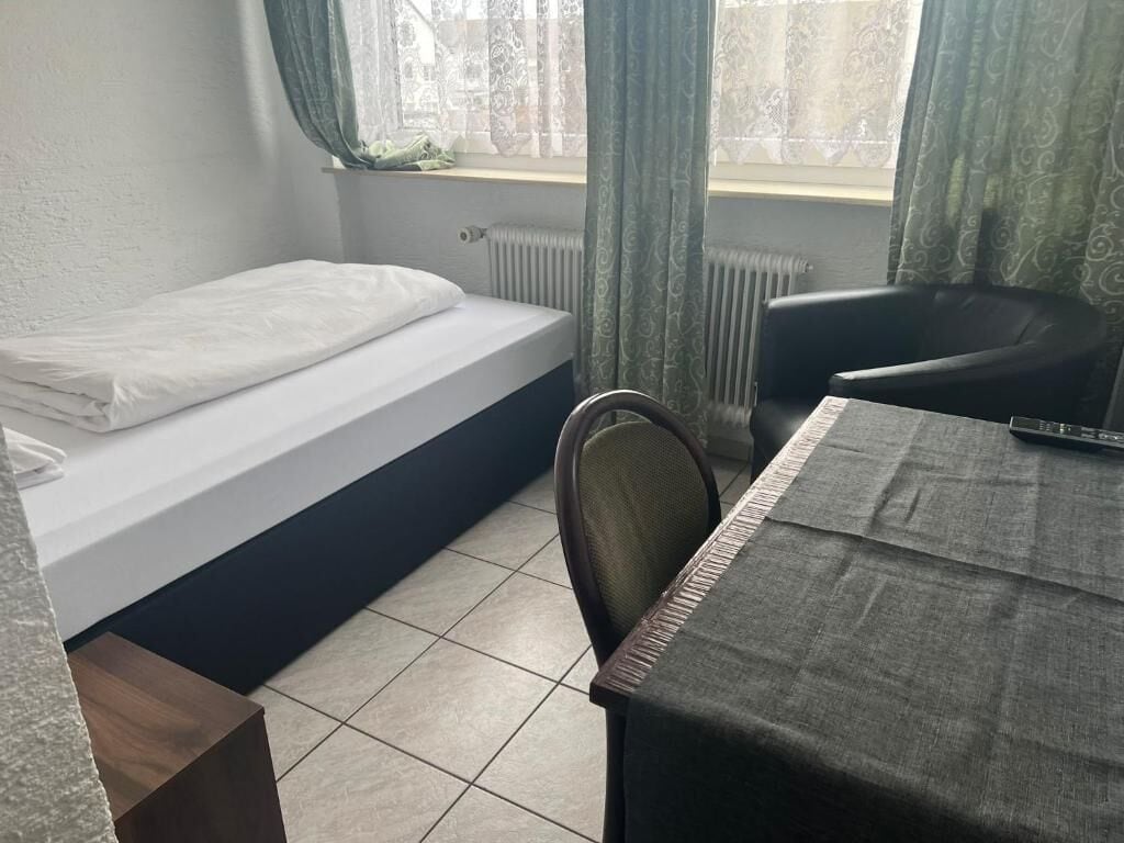 A small room with a bed, a table, and a chair. There is a window with curtains, a radiator, and a remote control on the table.