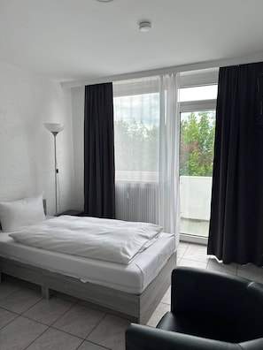 A hotel room with a bed, a chair, a floor lamp, and a view of trees through a window. - Hotel Main Taunus  (Hochheim am Main)
