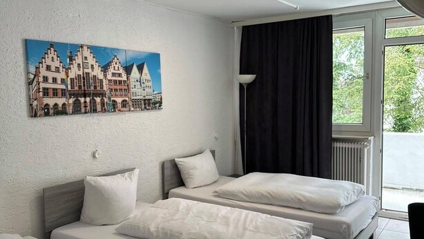 A hotel room with two beds, a painting of a European town, a window with curtains, and a radiator.