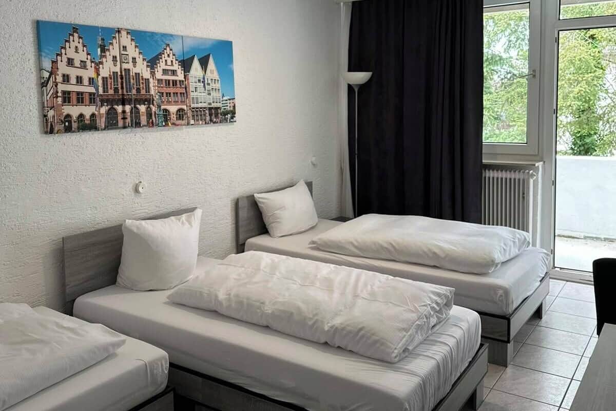 A hotel room with two beds, a painting of a European town, a window with curtains, and a radiator.
