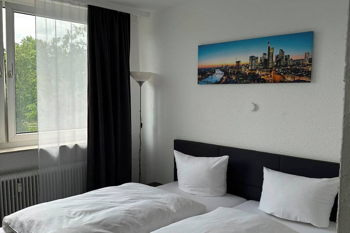 A hotel room with two beds, a cityscape painting, a window with curtains, and a radiator.