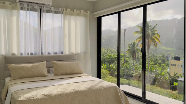 Une chambre à coucher avec un lit, des rideaux et une vue sur la verdure et les montagnes, grâce à de grandes fenêtres.