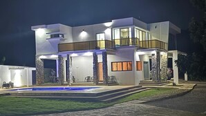 Une maison moderne à deux étages, dotée d’une piscine, de piliers en pierre et d’un éclairage extérieur soigné.
