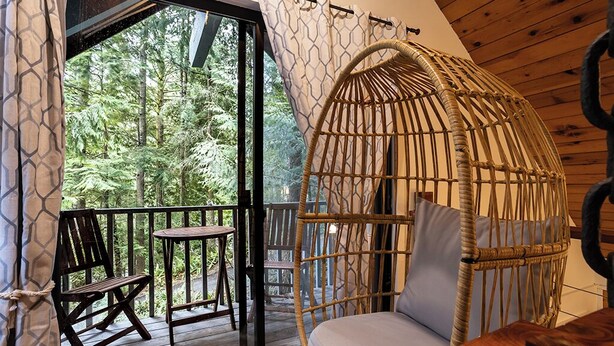 Um quarto aconchegante com uma cadeira de vime e assento acolchoado, uma mesa de madeira e vista para uma floresta através de uma porta de vidro.