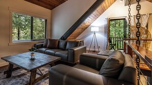 Uma sala de estar aconchegante no estilo loft, com teto de madeira, um conjunto de sofá, mesa de centro e uma grande janela com persianas.