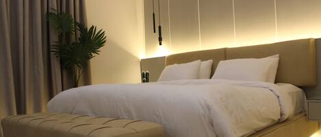 A modern bedroom with a large bed, a matching ottoman, a bedside table, and a potted plant.