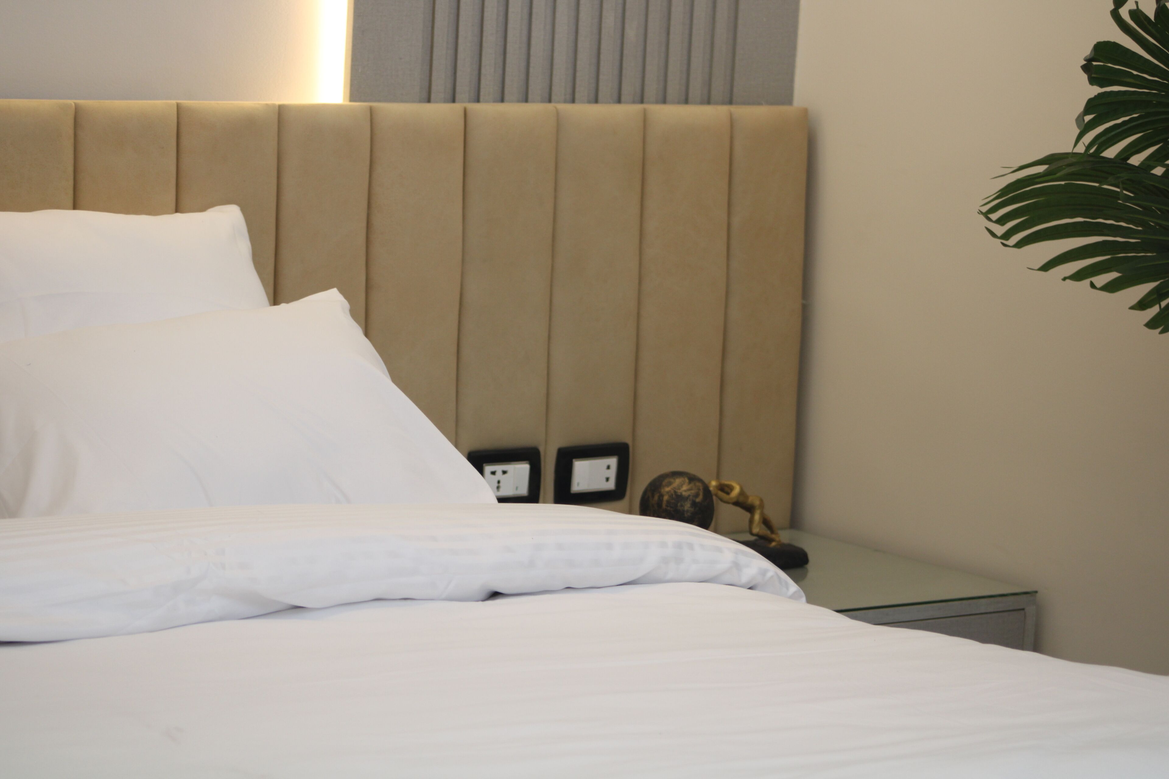 A neatly made bed with white linens, a beige upholstered headboard, and a small bedside table with a lamp and a decorative item.