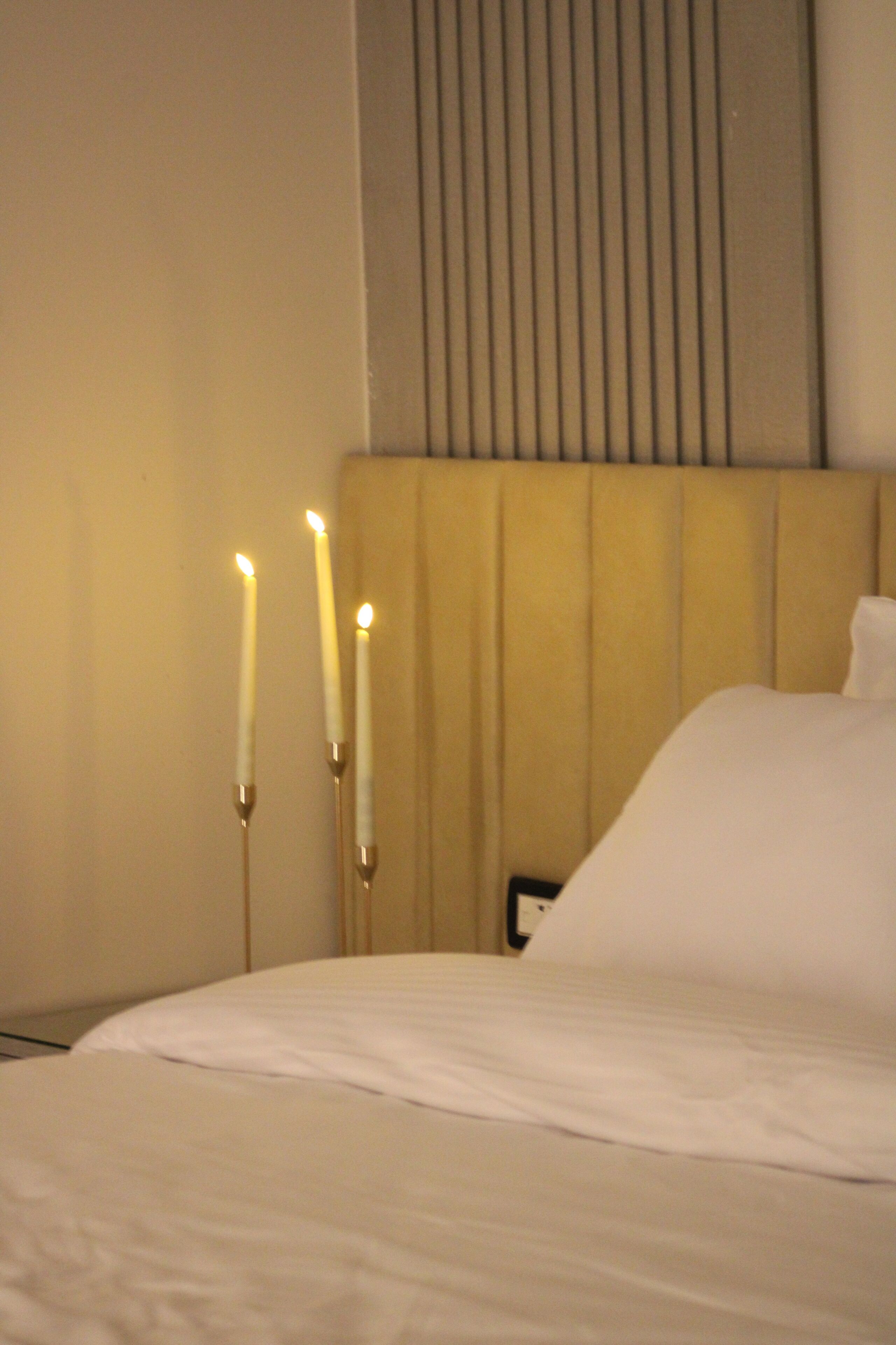A neatly made bed with white linens and three lit candles on a bedside table.