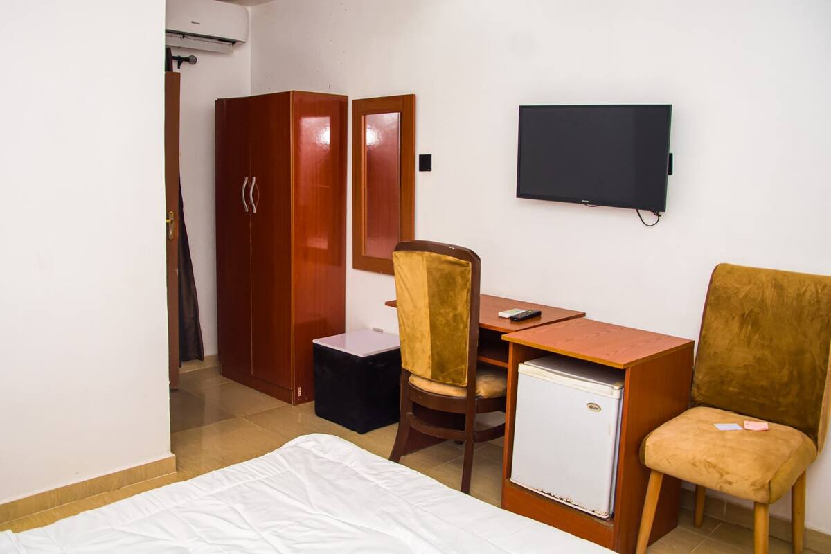 A hotel room with a bed, a desk with a chair, a small refrigerator, a wardrobe, and a television on the wall.