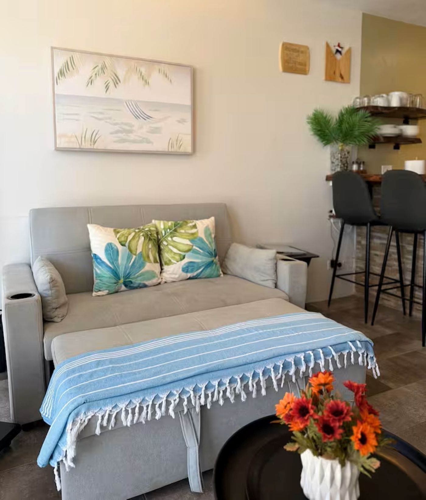 Ein Wohnzimmer mit einer Couch, einem Couchtisch mit blau-weiß gestreifter Tischläufer und einer Vase mit orangefarbenen Blumen.
