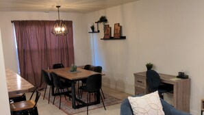 A dining area with a table and chairs, a blue armchair, a desk with a chair, and a hanging light fixture.