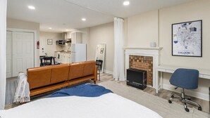 A modern hotel room with a bed, a desk, a chair, a fireplace, and a kitchen area in the background. - Cozy Studio Near Children’s Hospital, Kitchen & Laundry (Columbus)