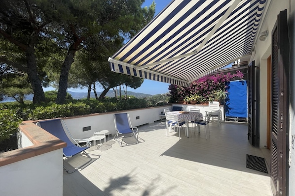 A covered outdoor seating area with chairs and tables, overlooking a body of water and trees. - Il Polpetto Apartment (Capoliveri)