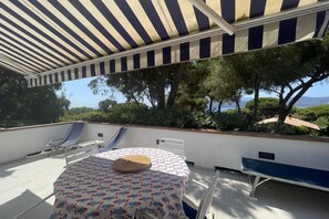 A covered outdoor seating area with a table and chairs, overlooking a scenic landscape with trees and a view of the sea.