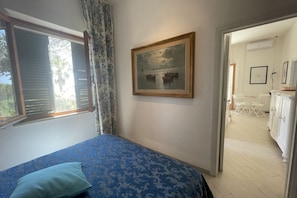 Un dormitorio con una cama, una ventana con contraventanas, un cuadro en la pared y vistas a otra habitación.