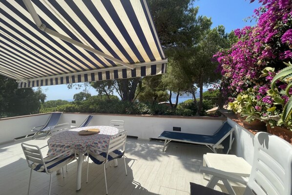A rooftop terrace with a striped awning, lounge chairs, a table with a patterned tablecloth, and a view of trees and flowers.