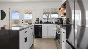 A modern kitchen with white cabinetry, a stainless steel refrigerator, and a view of the outdoors. - Joshua Tree Oasis • Cowboy Pool • Hot Tub • 5 Private Acres (Yucca Valley)
