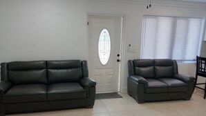 Ein Wohnzimmer mit zwei schwarzen Ledersofas, einer weißen Tür mit Fensterglas und einem Fenster mit Jalousien.