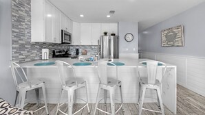 Una cocina moderna con armarios blancos, un refrigerador de acero inoxidable y un microondas. Hay dos taburetes de bar blancos con tapas turquesa.