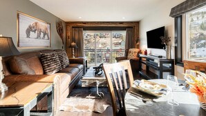 A cozy living room with a sofa, a coffee table, a dining table set for two, and a large window with a view of houses. - Rustic Luxe on Main | Spa, Dining & Charm On-Site at the Treasure Mountain Inn (Park City)