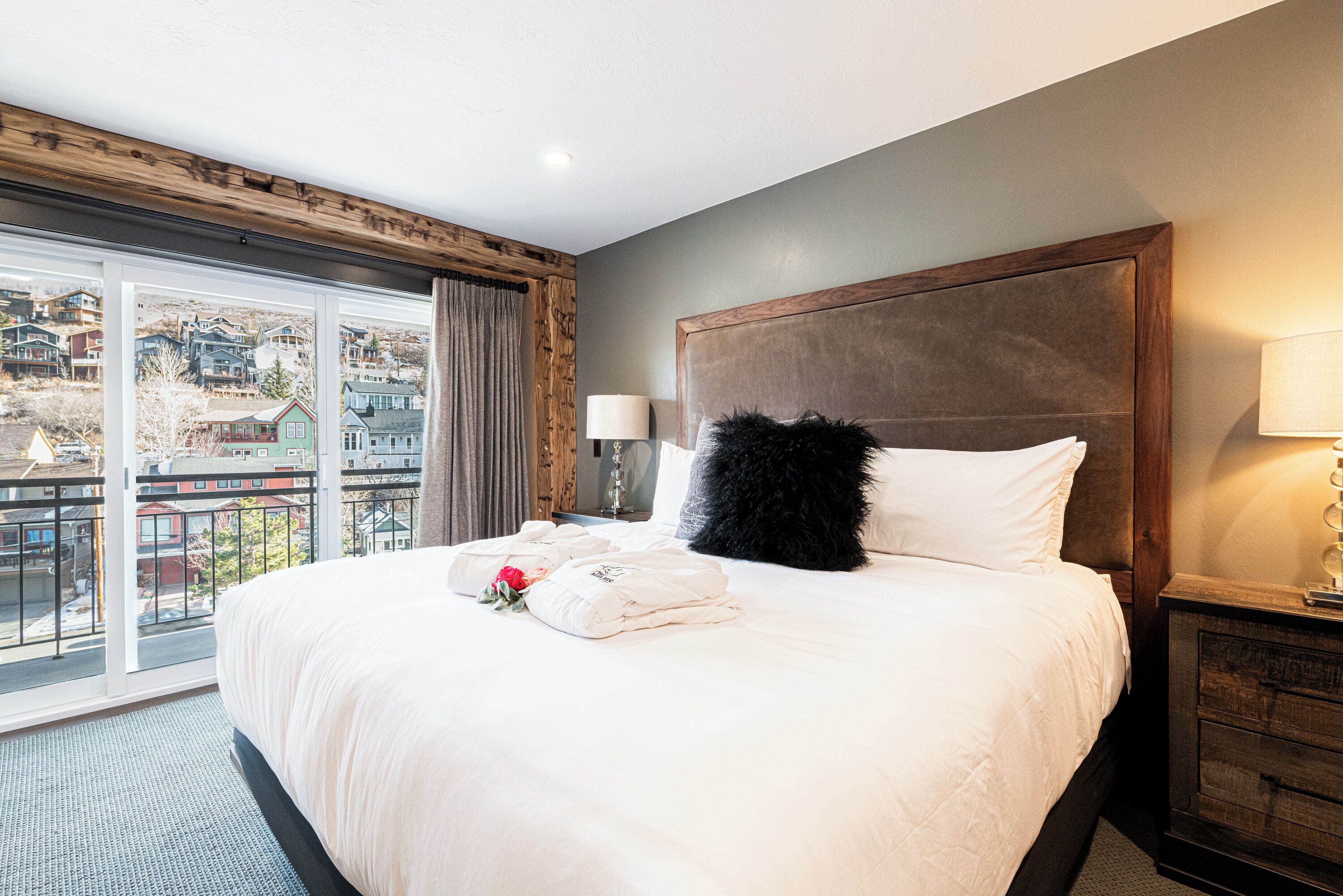 A hotel room with a large bed, a wooden headboard, a view of the city through large windows, and a balcony.