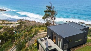 Une maison moderne avec un toit plat, de grandes fenêtres et un balcon donnant sur une plage et l’océan.