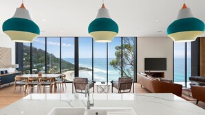 Un salon moderne doté d’une grande fenêtre donnant sur la mer, d’une cuisine avec un plan de travail en marbre et de trois suspensions lumineuses aux abat-jour turquoise et blancs.