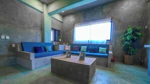 Een woonkamer met een blauwe bank, een bijzettafeltje, een raam met zonwering en een potplant.