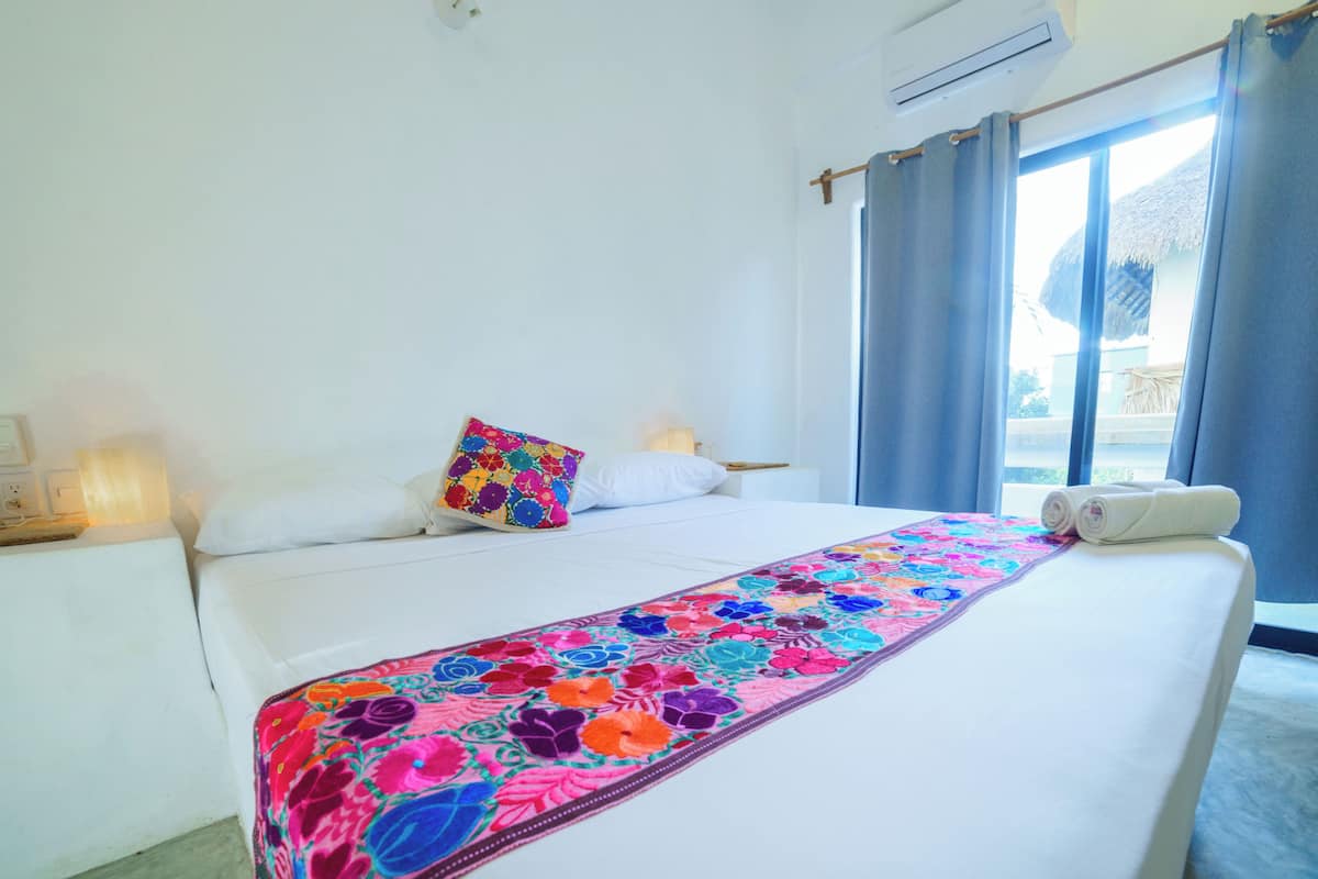 A hotel room with a bed, a colorful floral bedspread, a white pillow, a colorful pillow, a rolled towel, a nightstand with a lamp, and a window with curtains.