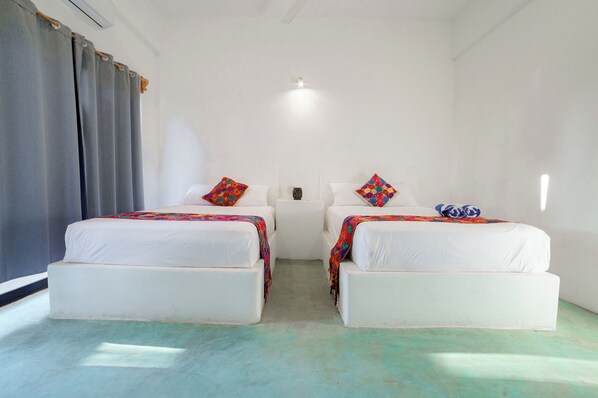 Una habitación con dos camas, cada una con una manta y almohada de colores, cabeceros blancos y una lámpara en el techo.
