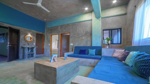 Een ruime woonkamer met een blauwe bank, een betonnen salontafel en een plafondventilator.
