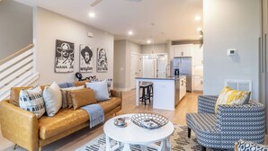 A modern living room with a sofa, coffee table, and patterned armchair, a kitchen with a fridge and island, and a staircase with white railings. - Monty by Avantstay 4bdr 4ba, Balcony w/ Outdoor Seating, 5mins to DT (Nashville)