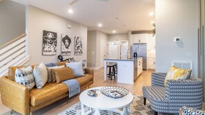 Sala de estar moderna com sofá, mesa de centro e poltrona estampada, cozinha com geladeira e ilha, e escada com corrimão branco.