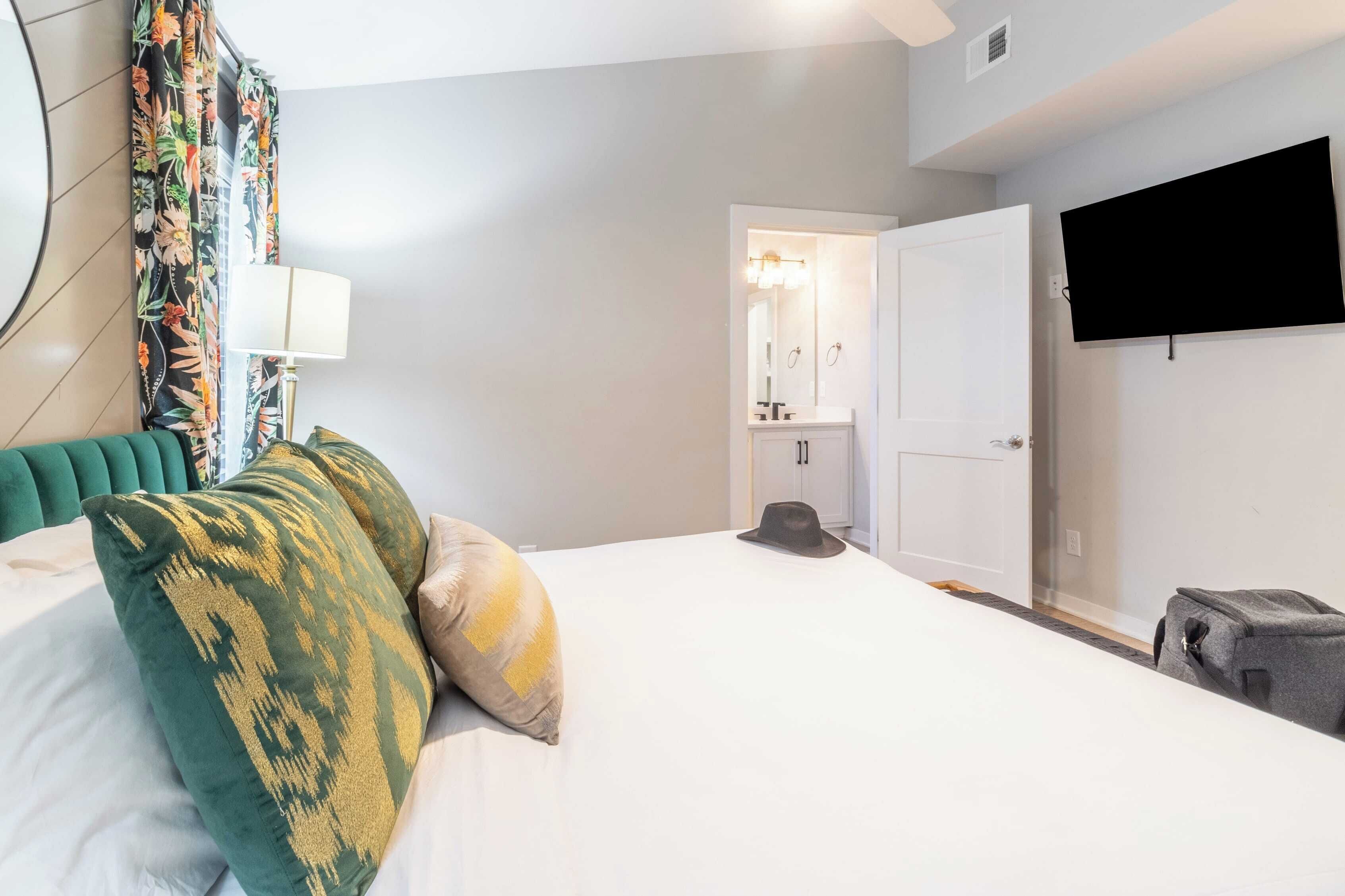 Una habitació d'hotel amb un llit, una televisió penjada a la paret, una porta que dona a un bany i una maleta a terra.