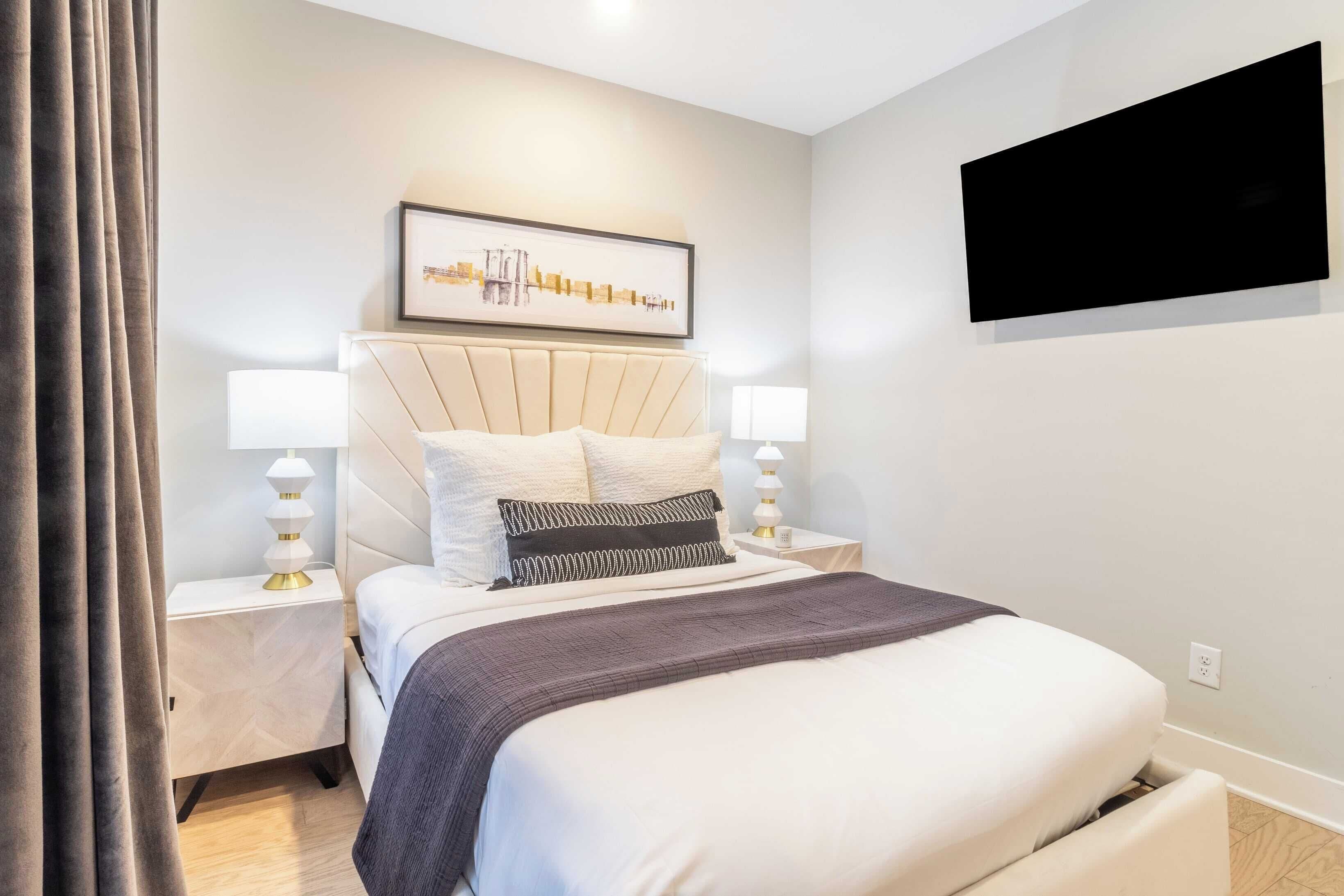 A modern hotel room with a large bed, a flat-screen TV mounted on the wall, and a framed artwork above the bed.