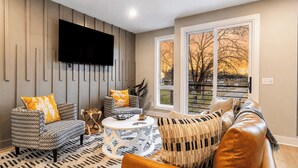 A living room with a TV mounted on the wall, a sofa, a coffee table, and a window with a view of trees. - Monty by Avantstay 4bdr 4ba, Balcony w/ Outdoor Seating, 5mins to DT (Nashville)