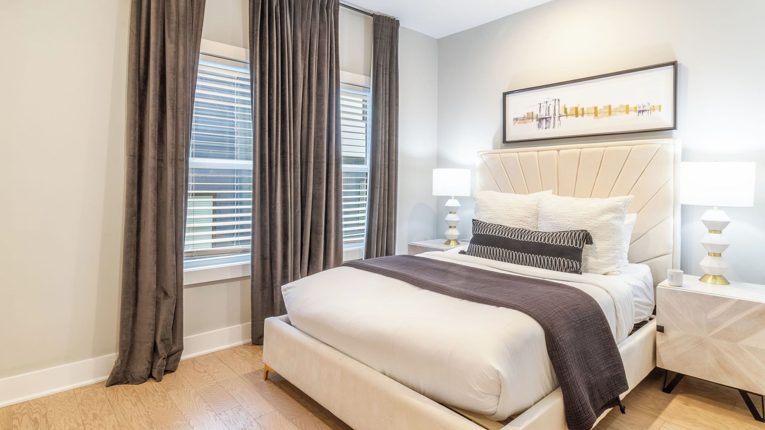 A bedroom with a bed, curtains, a headboard, bedside tables, and a wall-mounted artwork.